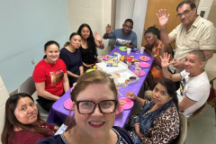 A group of smiling adults sitting around a rectangular table eating and enjoying  each others company.