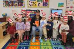 A group of smiling children on a carpet in a classroom wearing red and white Dr. Seuss hats; gathered around a male sitting on a chair holding a copy of the Dr. Seuss book, "Green Eggs and Ham"; all are looking straight towards the camera.