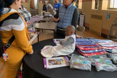 Three women standing  at a round table talking. The table is covered with baby supplies and a toy doll.
