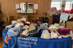 A smiling woman seated at a round table covered in a blue Catholic Charities table cloth with baby clothes on it.