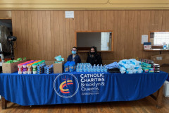 Two smiling women seated behind a long table covered with a blue Catholic Charities table cloth with a variety of baby products on it.