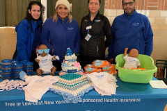 Four smiling individuals standing behind a table covered in a blue BlueCross BlueShield table cloth with baby items on it.