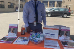 A man standing under a blue umbrella next to an table from Village Care Max.