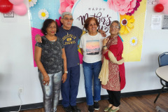 Three woman and one male are stand next to each other in front of a floral backdrop during a Mother's Day celebration .