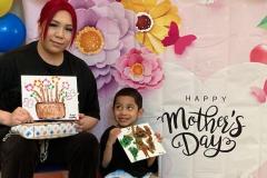 A woman and young boy hold floral paintings on canvas in front of a decorative Mother's Day backdrop.