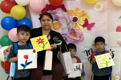 A woman and three young boys hold floral paintings on canvas in front of a decorative Mother's Day backdrop. surrounded by colorful balloons.