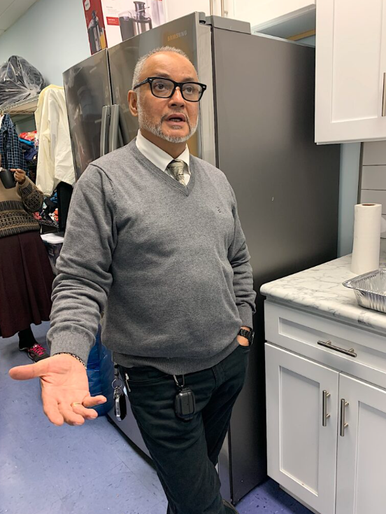 Man in a gray sweater and tie wearing glasses stands in a kitchen area speaking and making hand gestures.